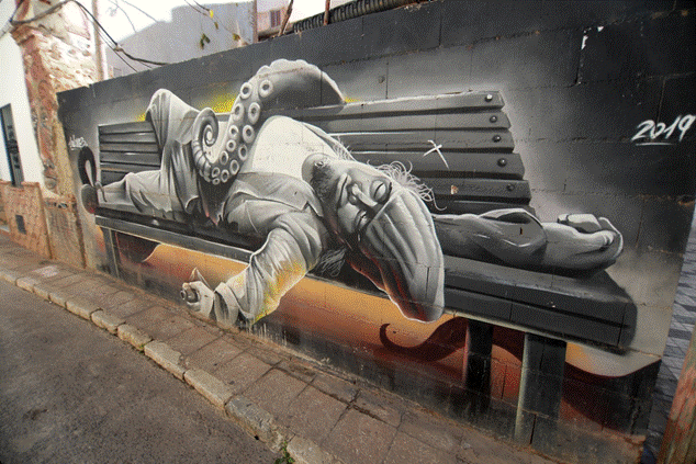 Mural urbano surrealista que representa una figura humana con cabeza de tentáculo de pulpo recostada en un banco, simbolizando la transformación y la identidad en el arte callejero.