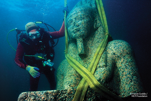 Un buceador junto a una gran estatua egipcia antigua bajo el agua durante una exploración arqueológica de una ciudad hundida.